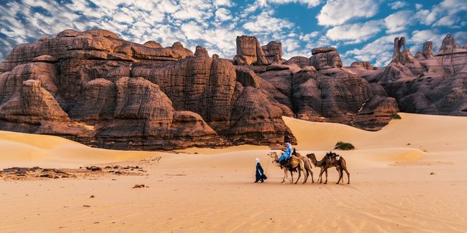 People wearing traditional robes ride camels in the desert in Algeria