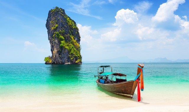 A wooden longtail boat on a white, sandy beach with a rock emerging from the turquoise sea nearby in Thailand