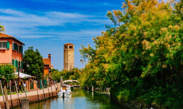 Una torre che s'intravede dietro un ponte che traversa un canale a Torcello, con alberi che sporgono ai lati
