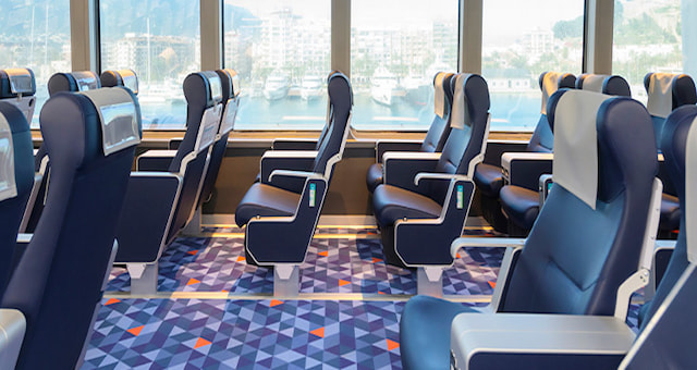 Interior view of a ferry cabin showing rows of modern, blue leather seats looking out at a harbor