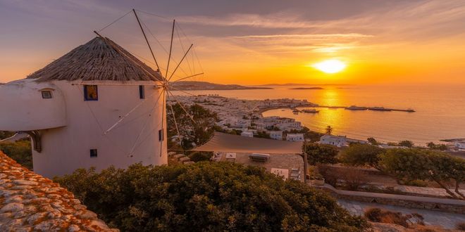 A traditional white Cycladic windmill against a colorful orange sunset sky in Mykonos, Greece