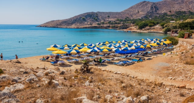 Sandy beach with rows of blue and yellow umbrellas and turquoise water in Halki, Greece