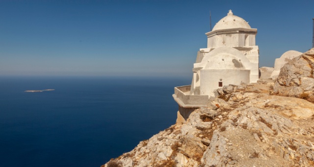 A white domed church perched on a steep rocky cliff overlooking the deep blue Aegean Sea in Anafi, Greece
