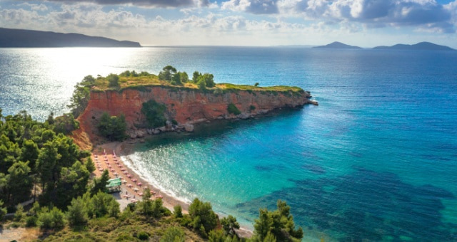 A red rocky cliff overlooking a sandy beach and clear blue sea with small islands in Alonissos, Greece